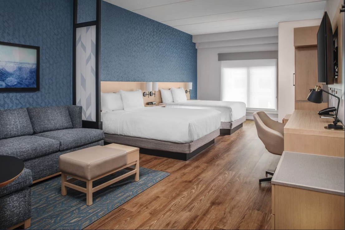 Modern hotel room with two beds, a sitting area, desk, and a large window, featuring a blue accent wall and wooden flooring.